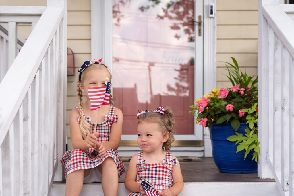 Two little girls sitting on the front porch of a charming home waving American flags